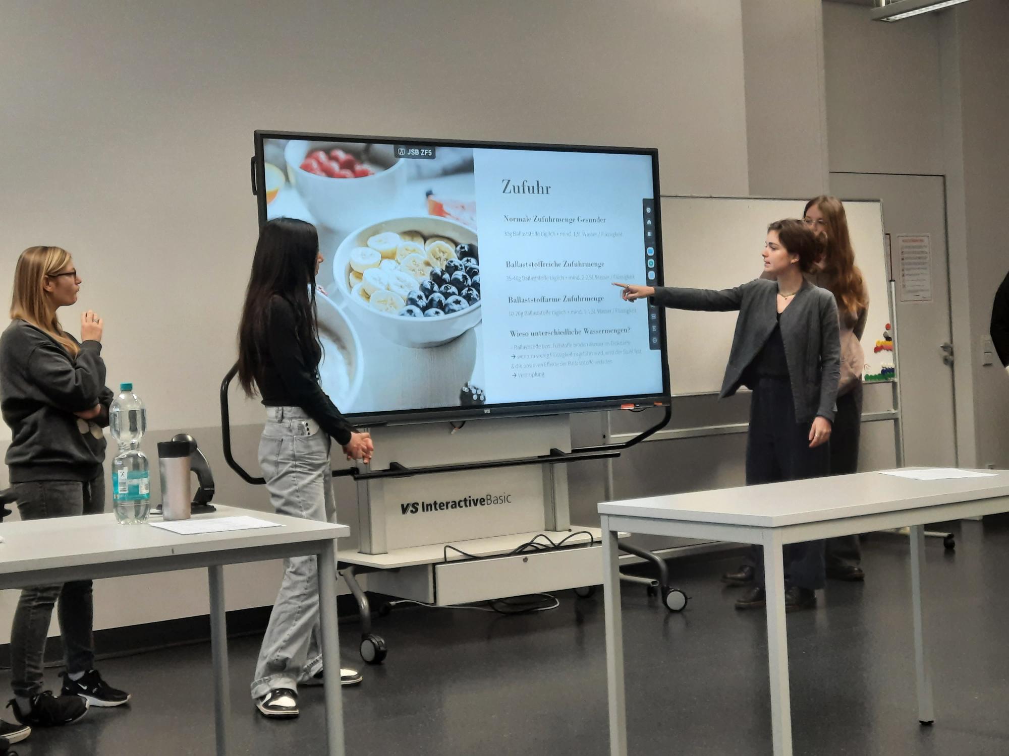 Vier Personen in einem Klassenzimmer, eine Frau zeigt auf einen Bildschirm mit einer Diätpräsentation.