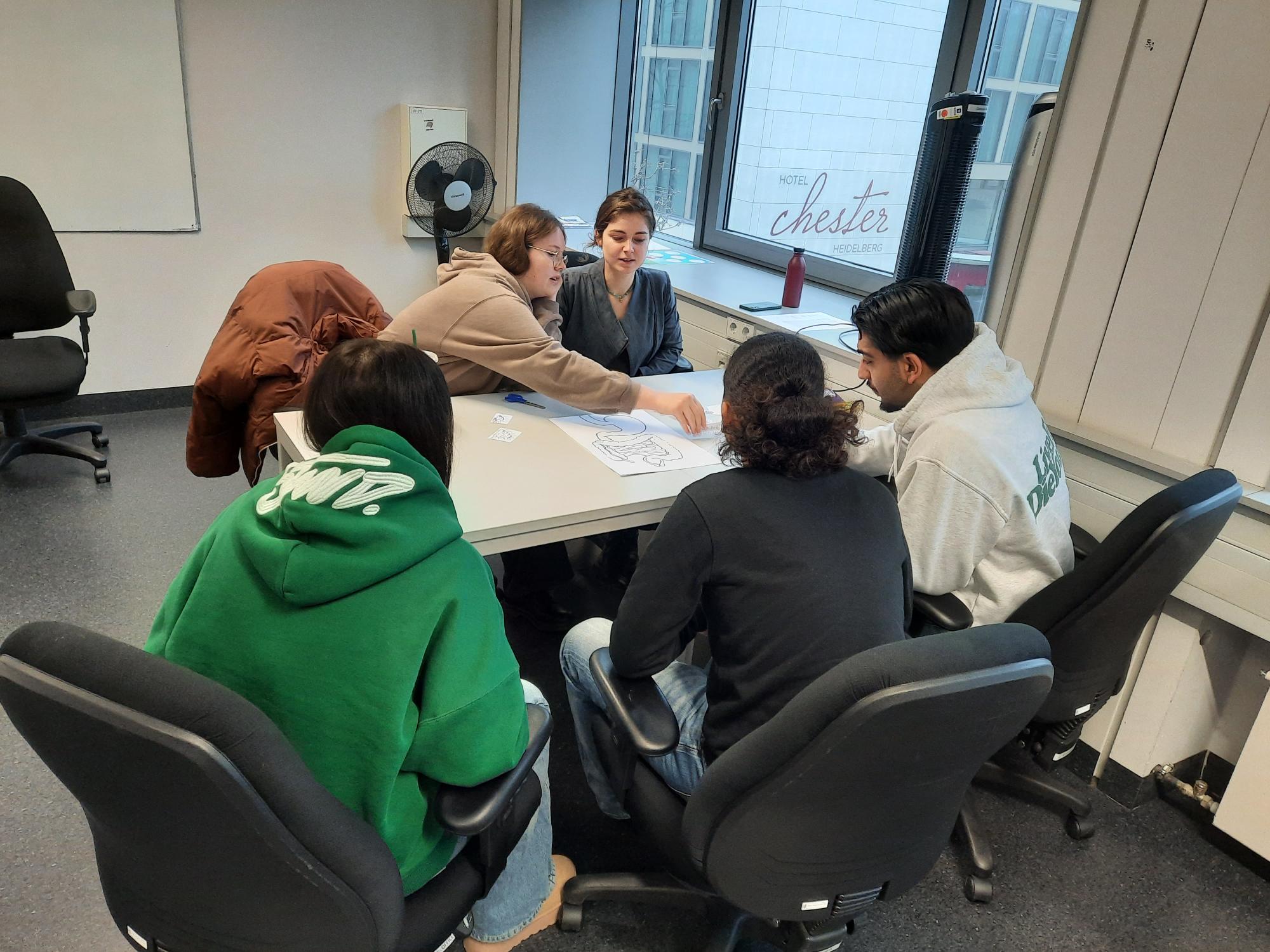 Eine Gruppe von fünf Personen sitzt um einen Tisch und arbeitet an einem Projekt.