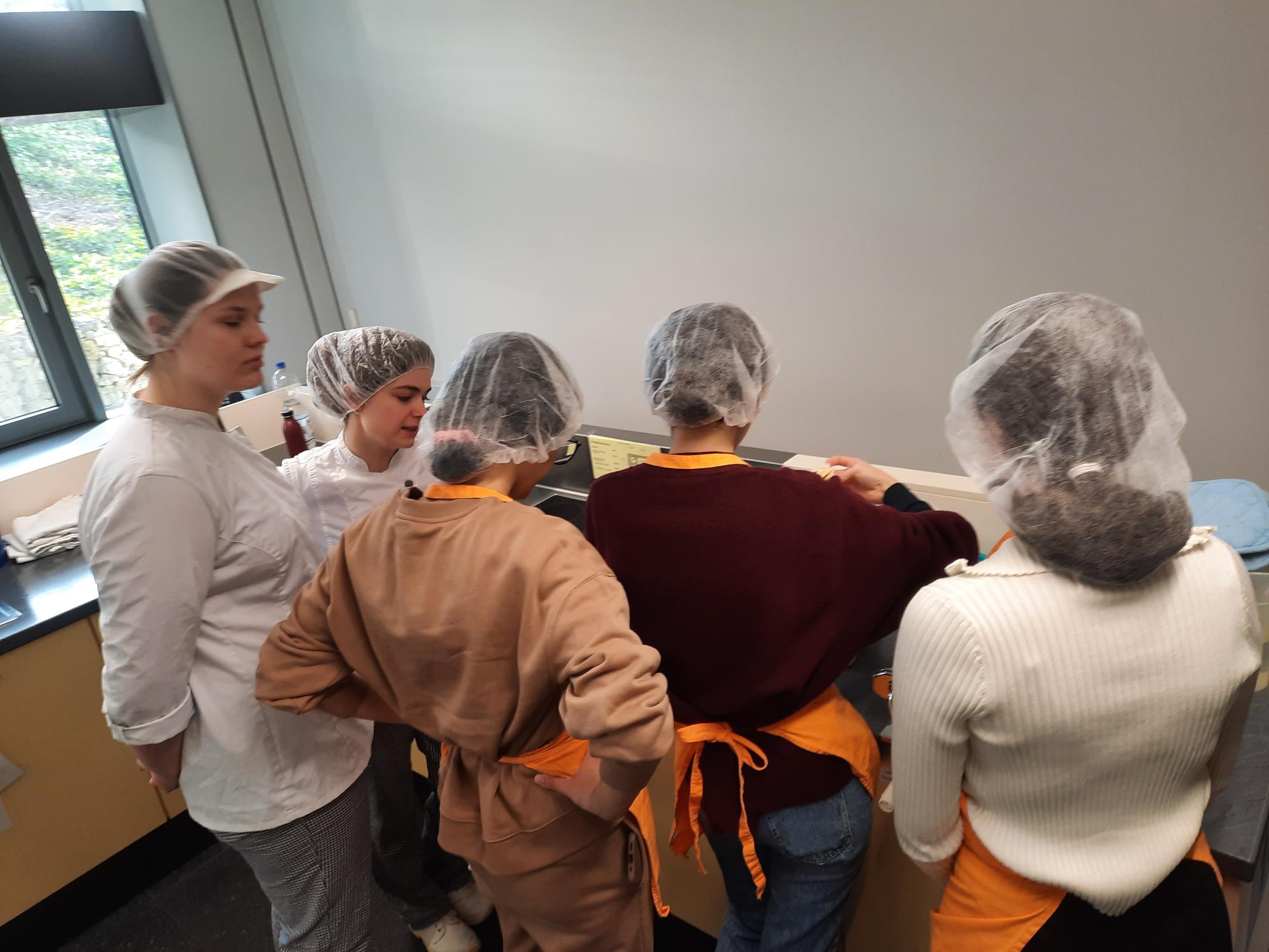 Eine Gruppe von Menschen in Schürzen und Hauben steht um einen Tisch.