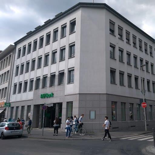 Ein mehrstöckiges Gebäude mit einem grünen Schild, Menschen und Fahrrädern vor dem Gebäude.