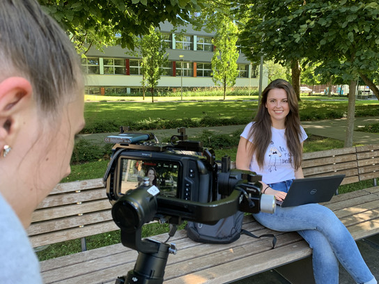 Eine Frau sitzt auf einer Bank und arbeitet an einem Laptop, während eine andere Person eine Kamera auf sie richtet.