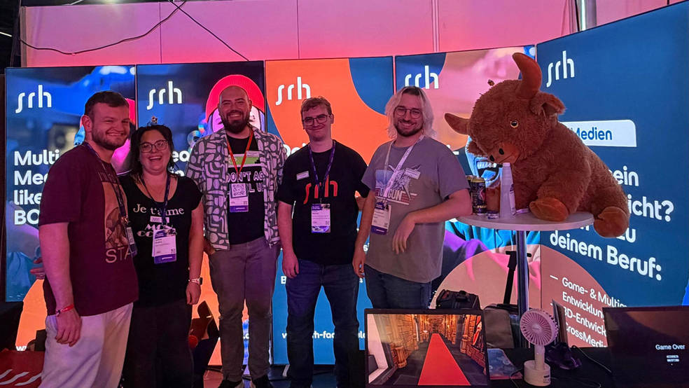 Gruppenfoto des fünfköpfigen SRH-Teams vor den leuchtenden Bannern des Messestands auf der gamescom. Rechts sitzt ein großes Plüschtier in Form eines Hochlandrinds auf einem Stehtisch, vorne läuft auf einem Bildschirm ein selbst entwickeltes Spiel.