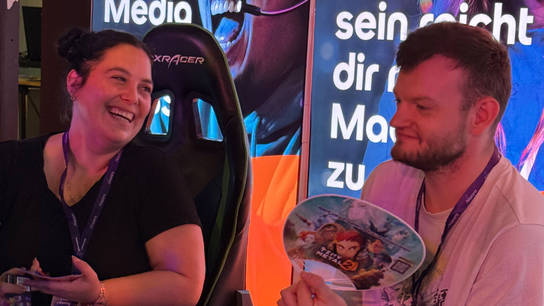 Zwei Teammitglieder der SRH lachen und unterhalten sich am Messestand, im Hintergrund ein Gaming-Stuhl und beleuchtete Werbebanner. Einer der beiden hält einen Fächer mit Spielmotiv in der Hand.