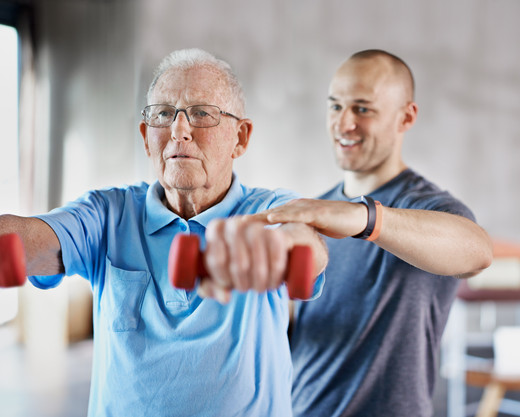Physiotherapeut gibt älterem Herrn bei einer Übung mit Hanteln Hilfestellung
