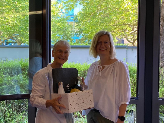 Zwei Frauen stehen vor einem Fenster, eine hält eine Geschenkbox.