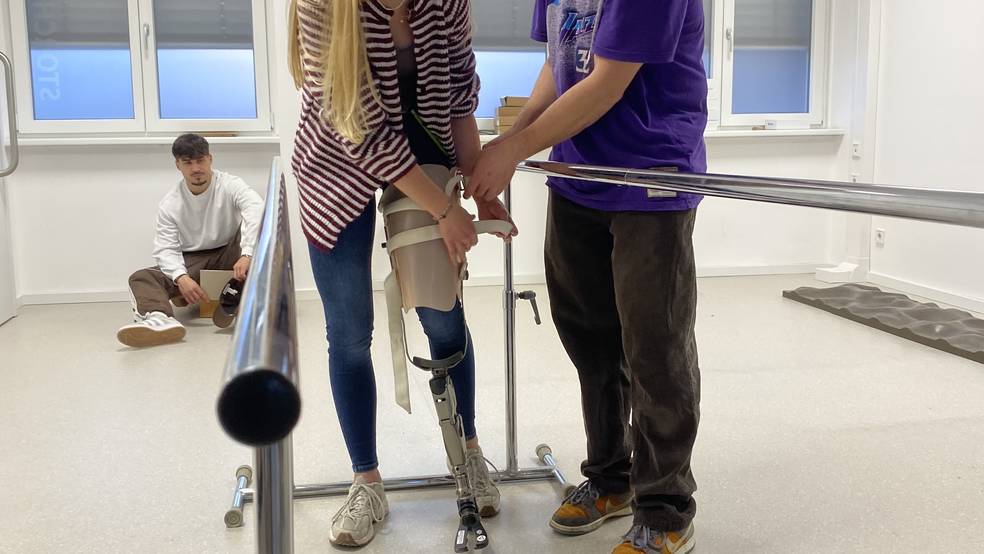 In einem hellen Therapieraum übt eine junge Frau das Gehen mit einer Beinprothese zwischen zwei Parallelbarren. Ein Mann steht unterstützend neben ihr und hilft bei der Anpassung und Stabilisierung der Prothese. Beide lächeln in die Kamera. Im Hintergrund sitzt eine weitere Person auf dem Boden und beobachtet die Situation. Die Szene vermittelt eine praxisnahe Übung aus dem Bereich Orthopädietechnik und Rehabilitation.