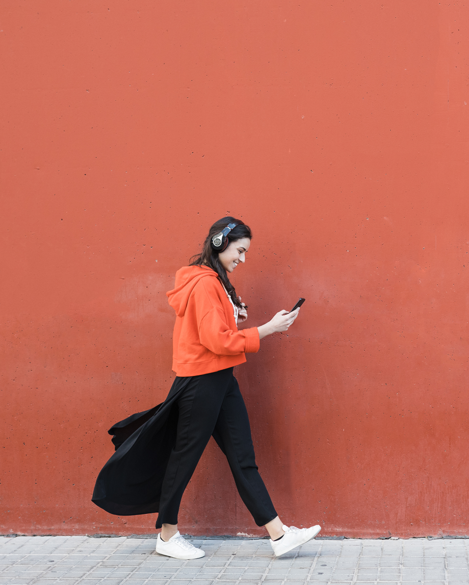 Mädchen vor orangenem Hintergrund Eine Person in einem orangen Kapuzenpullover geht an einer roten Wand und schaut auf ihr Handy.