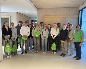 Eine Gruppe von etwa 18 jungen Erwachsenen und mehreren Lehrkräften steht nebeneinander in einem hellen, modernen Innenraum. Einige halten grüne Stoffbeutel in der Hand. Die Gruppe blickt lächelnd in die Kamera; im Hintergrund sind helle Holzwände und ein Flur zu sehen.