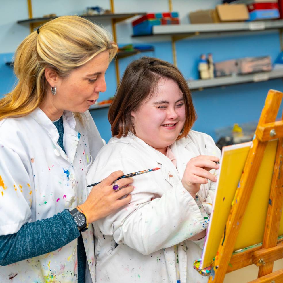 Ergotherapeutin schaut einer Frau mit Downsyndrom beim Malen auf einer Leinwand zu