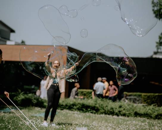 Eine Person bläst große Seifenblasen auf einem grasbewachsenen Bereich, während andere im Hintergrund stehen.