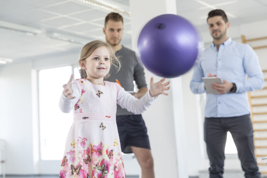 Ein kleines Mädchen in einem Blumenkleid spielt mit einem lila Ball, während zwei Männer im Hintergrund zuschauen.