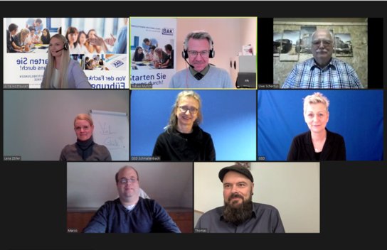 Ein Bild einer Videokonferenz mit neun Teilnehmern in einem Raster, die alle lächeln und in verschiedene Richtungen schauen.