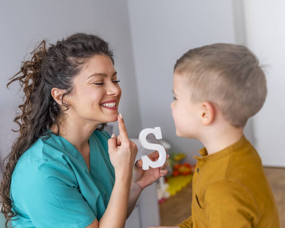 Sprachtherapiesitzung mit einem kleinen Jungen: Logopädin hält den Buchstaben Klotz S in der Hand und zeigt auf ihre Lippen für die Aussprache.