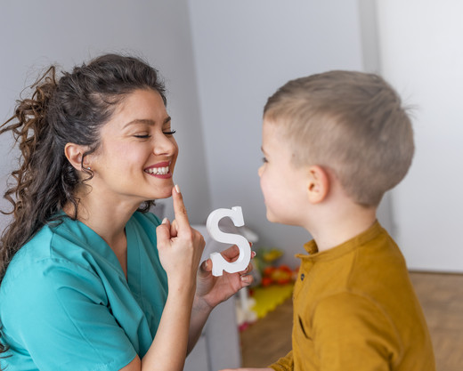 Sprachtherapiesitzung mit einem kleinen Jungen: Logopädin hält den Buchstaben Klotz S in der Hand und zeigt auf ihre Lippen für die Aussprache.