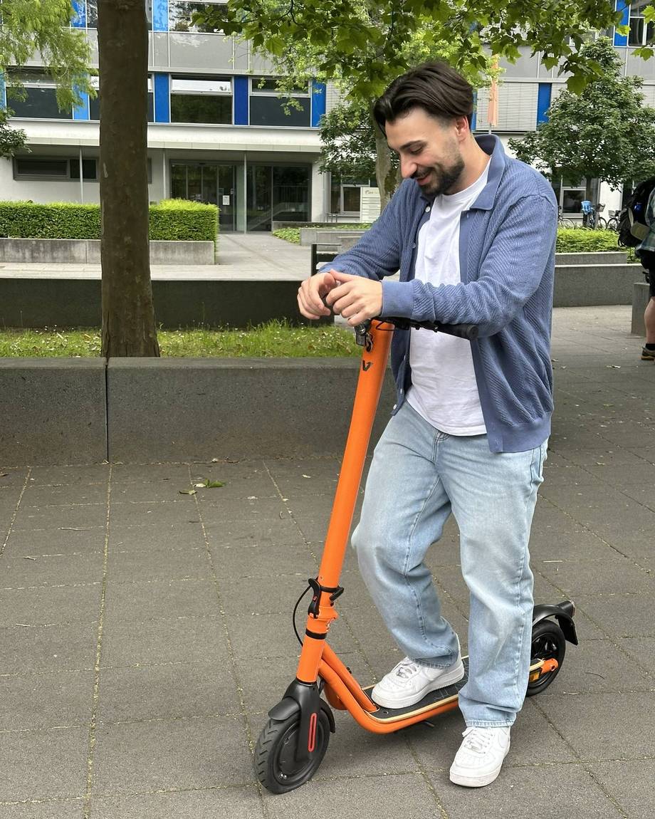Ein Mann schiebt ein orangefarbenes E-Scooter auf einem Gehweg.