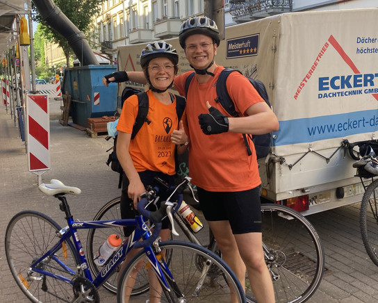 Zwei Personen in orangefarbenen T-Shirts und Fahrradhelmen stehen mit Fahrrädern vor einem weißen LKW.