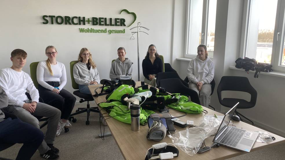 In einem hellen Besprechungsraum des Sanitätshauses Storch und Beller sitzen mehrere Studierende auf Bürostühlen um einen Tisch. Auf dem Tisch liegen grüne Stoffbeutel sowie orthopädietechnische Hilfsmittel und Geräte zur Muskelstimulation. An der Wand ist das Logo „STORCH + BELLER – Wohltuend gesund“ zu sehen. Die Teilnehmenden blicken aufmerksam in die Kamera, die Atmosphäre wirkt ruhig und professionell.