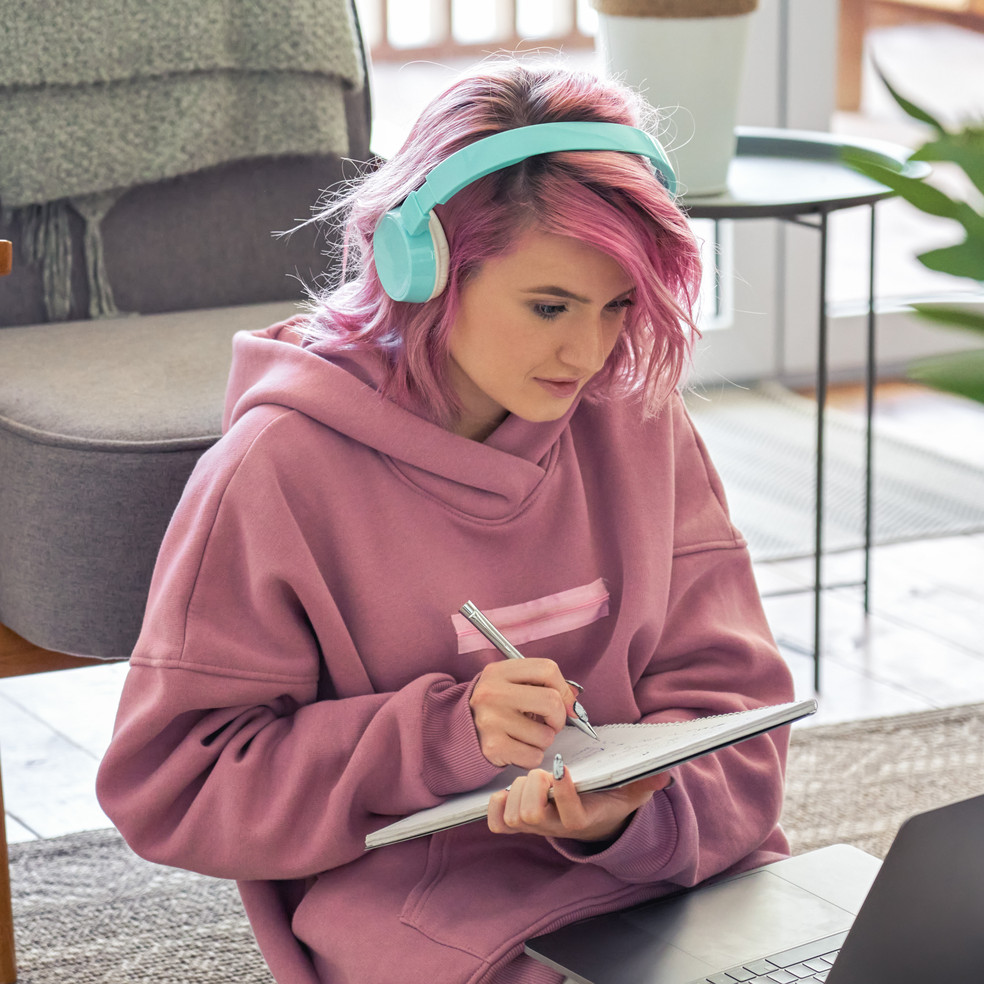 Pinkhaarige junge Frau sitzt auf dem Boden mit Laptop auf den Knien und Kopfhörern auf dem Kopf