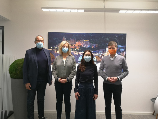 Vier Personen stehen vor einem Bild einer Stadtlandschaft, alle tragen Masken.