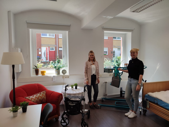 Zwei Frauen in einem Raum mit einem Rollator und einer Pflanze.