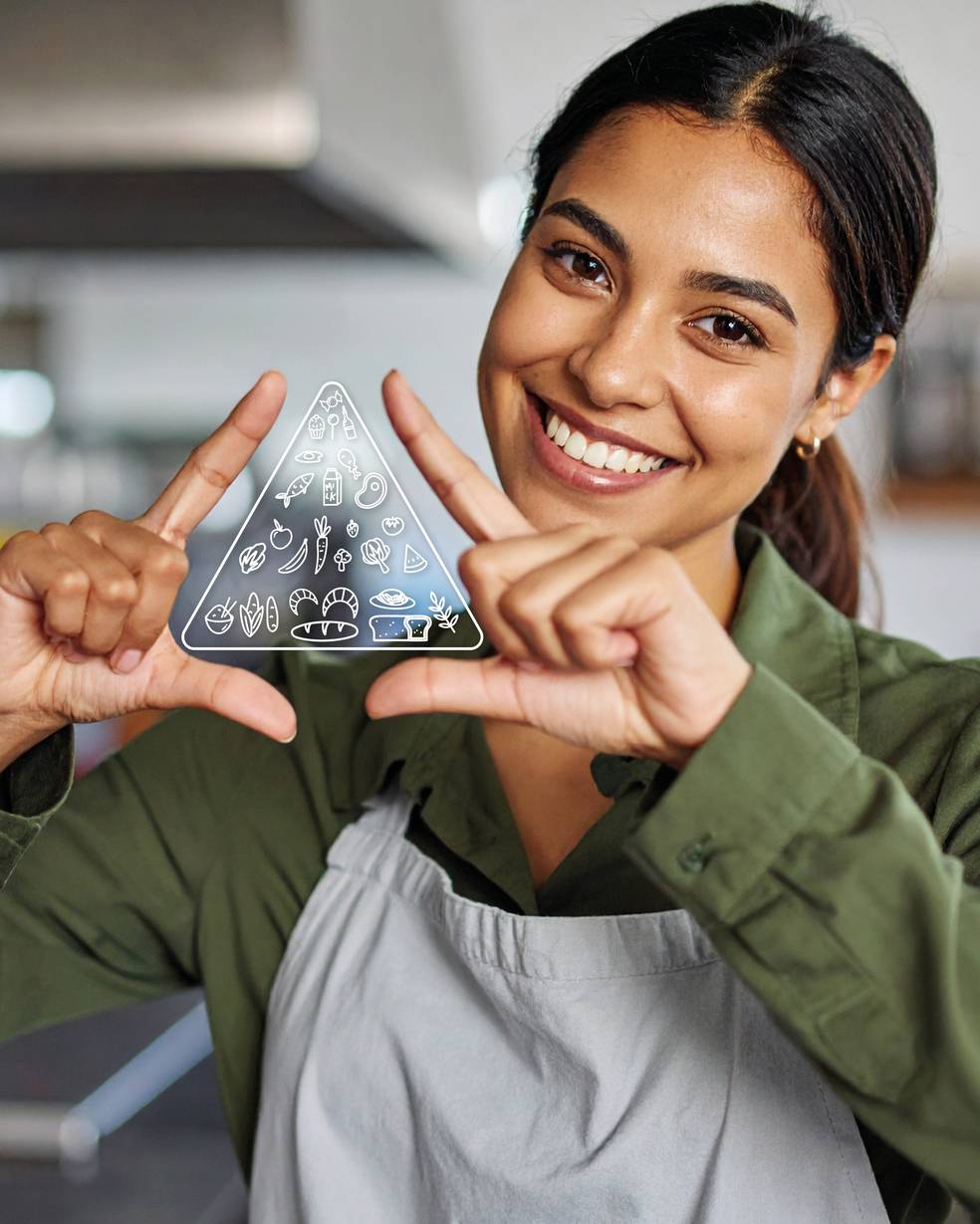 Eine Person in Kochjacke und grüner Schürze hält ein dreieckiges Symbol mit verschiedenen Lebensmittel-Icons. Sie lächelt und zeigt mit beiden Händen auf das Symbol.