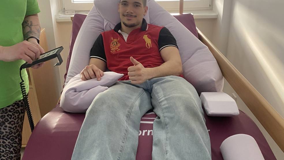 Bildbeschreibung / Alternativtext: Ein junger Mann liegt entspannt auf einer speziellen Therapieliege in einem Behandlungsraum und zeigt lächelnd einen Daumen nach oben. Über ihm befindet sich ein Haltegriff zur Unterstützung von Lagerung oder Transfer. Links steht eine Betreuungsperson in grüner Kleidung und bedient ein medizinisches Steuergerät. Die Szene vermittelt eine praktische Demonstration aus dem Bereich Pflege, Therapie oder Hilfsmittelversorgung.