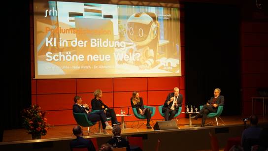 Eine Gruppe von Menschen sitzt auf einer Bühne vor einem großen Bildschirm mit dem Text 'KI in der Bildung - Schöne neue Welt?