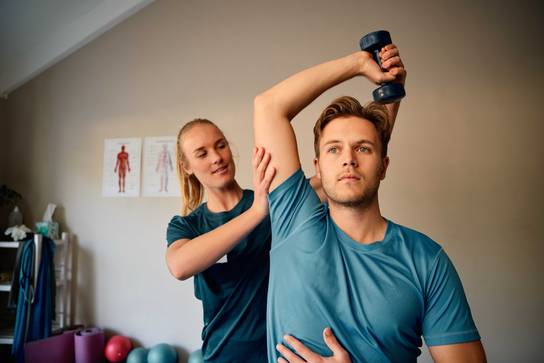 Foto Physiotherapie Ein Mann bei der Physiotherapie trainiert mit Hanteln, während eine Frau seine Haltung korrigiert.