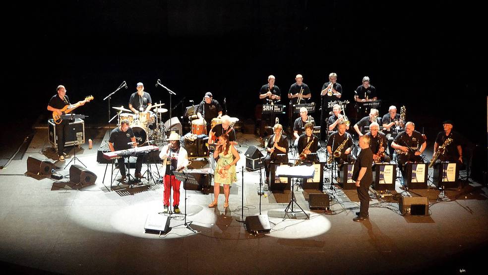 Eine große Band spielt auf einer Bühne, mit Musikern, die verschiedene Instrumente spielen.