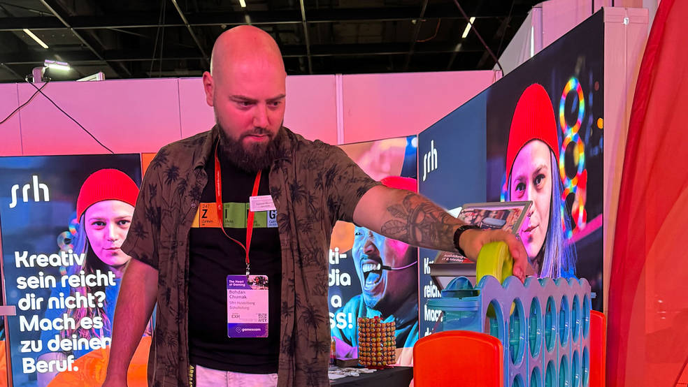 Ein Mitarbeiter der SRH Heidelberg spielt am Messestand eine Runde 4-Gewinnt und wirft gerade eine Spielscheibe ein. Im Hintergrund sind die farbigen SRH-Banner mit dem Slogan "Kreativ sein reicht dir nicht? Mach es zu deinem Beruf." zu sehen.