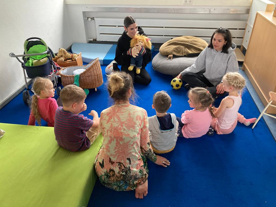 Eine Gruppe von Kindern sitzt auf einem blauen Teppich in einem Raum, umgeben von Spielzeugen und zwei Erwachsenen.