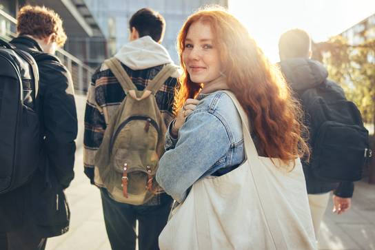 Eine junge Frau mit rotem Haar, die eine weiße Tasche trägt, steht in einer Gruppe von Menschen.