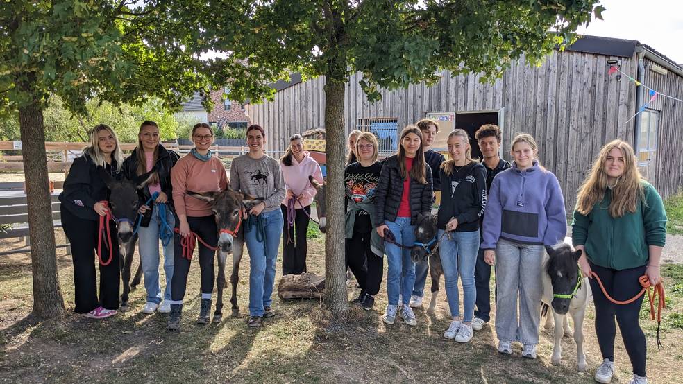 Ein Gruppenfoto zeigt etwa 15 junge Logopäd:innen, die in einer Reihe vor einer hölzernen Scheune auf einem Begegnungshof stehen. Sie haben an verschiedenen Stellen mit Tieren interagiert und halten jetzt die Leinen von vier Eseln und einem Mini-Shetlandpony in der Hand. Die Gruppe lächelt in die Kamera und wirkt entspannt. Zwischen ihnen ist ein junger Baum.