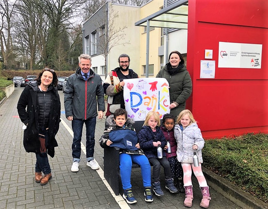 Eine Gruppe von Erwachsenen und Kindern posiert vor einem roten Gebäude, einige halten ein Schild mit der Aufschrift 'Danke'.