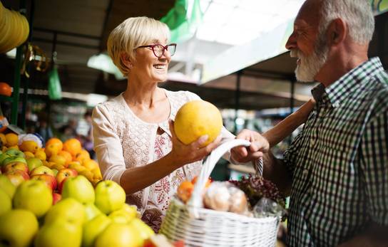 Ein älteres Paar lacht an einem Obststand, während die Frau eine Zitrone hält.